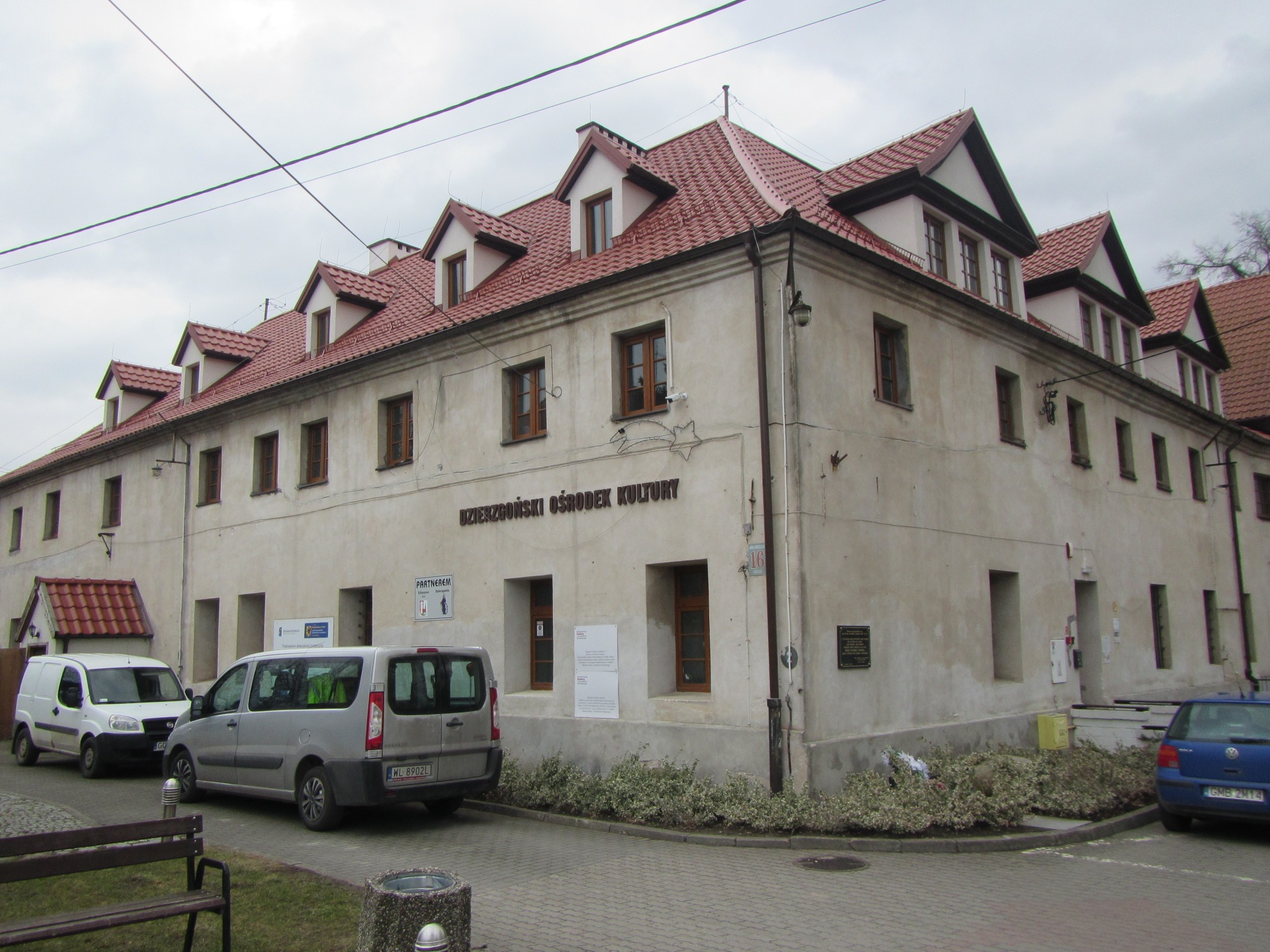 Budynek Dzierzgońskiego Ośrodka Kultury z czerwonym dachem i kilkoma oknami, zaparkowane samochody dostawcze przed budynkiem.