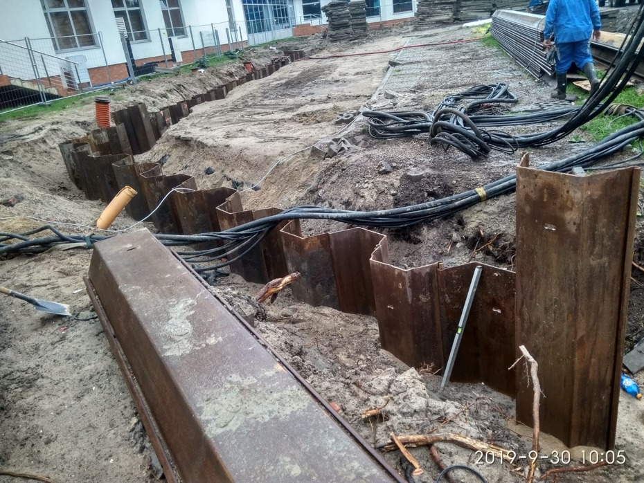 Wykop zabezpieczony stalowymi szalunkami, widoczne kable energetyczne, rury kanalizacyjne i poziomica, teren budowy w trakcie prac ziemnych.