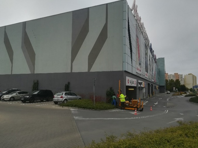 Prace geotechniczne przy parkingu centrum handlowego: żółta wiertnica w akcji, pracownicy w odblaskowych kamizelkach, białe pachołki ostrzegawcze.