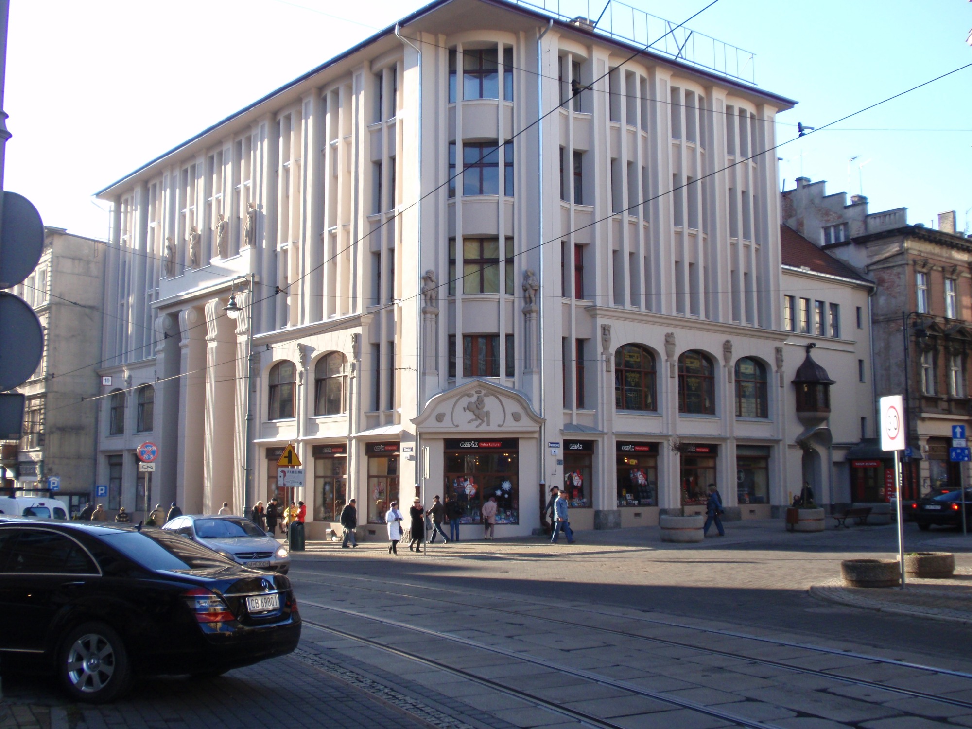 Kamienica w jasnej kolorystyce z rzeźbami na fasadzie, widoczna z perspektywy ulicy z tramwajami i przechodniami.