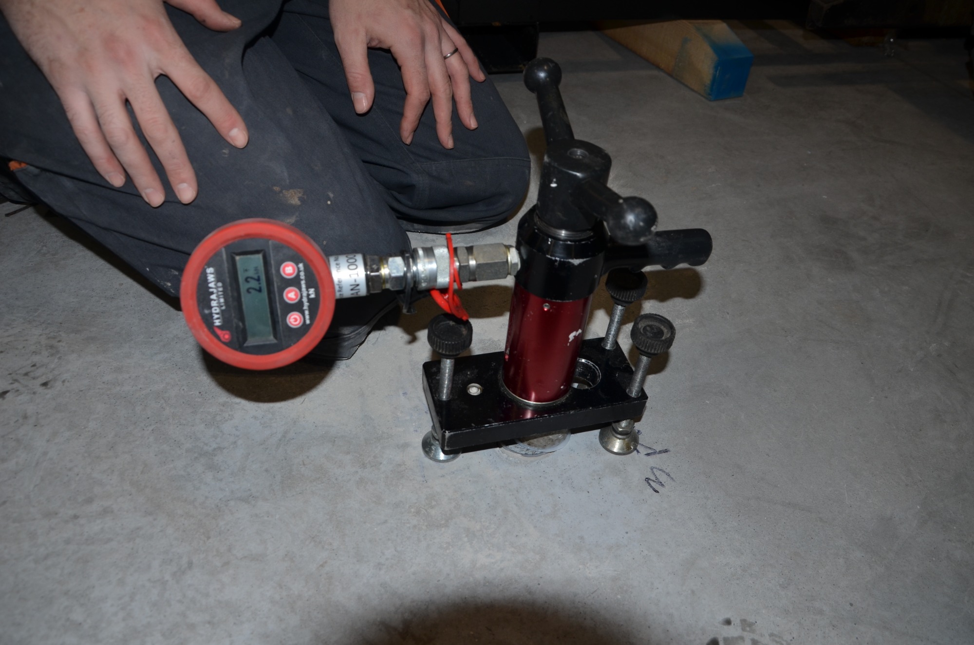 Urządzenie hydrauliczne z manometrem cyfrowym, używane do badania wytrzymałości betonu na ściskanie, ustawione na szarej powierzchni betonowej, obsługiwane przez osobę w ciemnych spodniach.