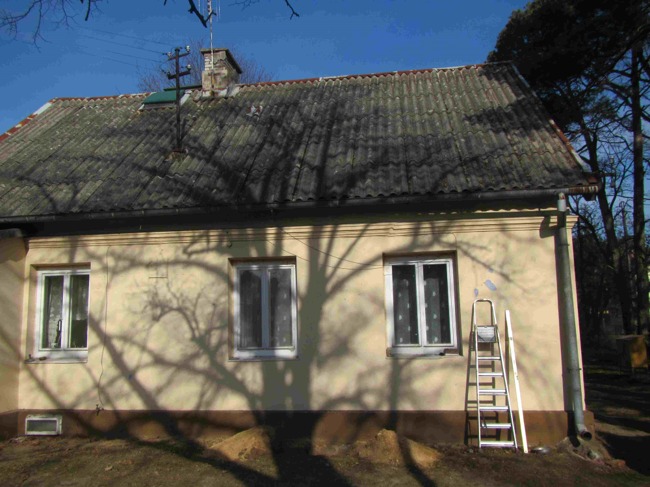 Dom jednorodzinny z szarym, falistym dachem, widoczne trzy okna z białymi ramami, ściana w kolorze kremowym z plamami, cień drzewa na elewacji, drabina oparta o ścianę, rynna spustowa.