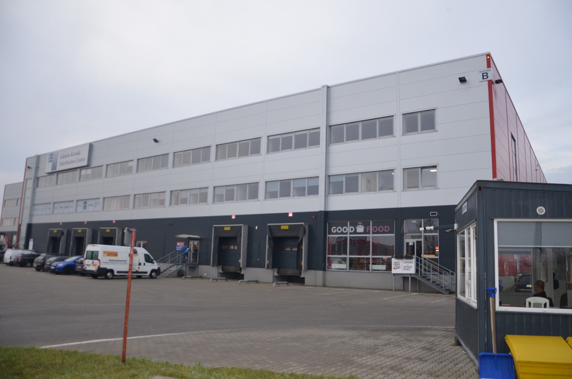 Szeroki kąt na budynek magazynowy z rampami załadunkowymi, napisem 'Gdańsk Kowale Distribution Centre' i budką strażniczą z niebieską łopatą i żółtymi pojemnikami z przodu.