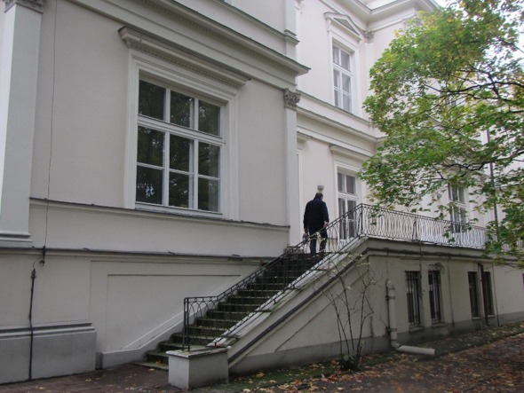 Elewacja budynku z balkonem i schodami zewnętrznymi, osoba w ciemnym płaszczu wchodzi po schodach.