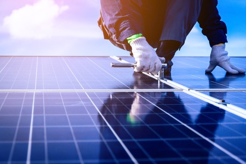 Instalacja paneli słonecznych na dachu: monter w rękawicach dokręca śruby mocujące konstrukcję wsporczą. Widoczne niebo i odbicia w panelach.