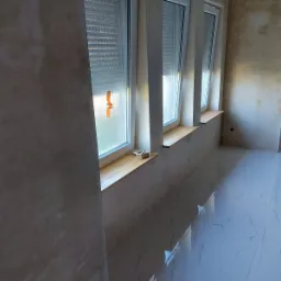 Wnętrze pokoju w trakcie prac wykończeniowych: ściany po nałożeniu tynku, widoczne trzy okna z roletami, białe płytki na podłodze i drabina malarska.