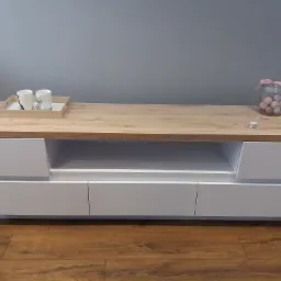 Nowoczesna szafka RTV z białymi frontami i blatem w odcieniu jasnego drewna, na blacie dekoracje: dwie filiżanki na tacy, szklany słoik z pastelowymi kulkami oraz mały świecznik.