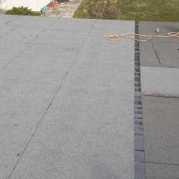 AlpinZerg Sp.z o.o - Widok z góry na płaski dach pokryty papą, częściowo świeżo położoną, z widoczną butlą gazową i narzędziami dekarskimi, sugerującymi prace naprawcze lub wykończeniowe.