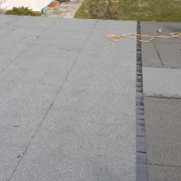 Widok z góry na płaski dach pokryty papą, częściowo świeżo położoną, z widoczną butlą gazową i narzędziami dekarskimi, sugerującymi prace naprawcze lub wykończeniowe.