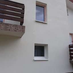 AlpinZerg Sp.z o.o - Świeżo otynkowana elewacja domu jednorodzinnego w jasnym odcieniu, z widocznymi oknami zabezpieczonymi folią malarską i balkonem z drewnianą balustradą oraz widocznymi wiadrami i narzędziami...