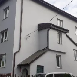 Wielopiętrowy budynek mieszkalny z szarą elewacją, brązowymi elementami dachu i rynnami, częściowo zasłonięty przez sąsiedni budynek z czerwonym dachem i drewniany płot, widoczne rusztowanie przy...