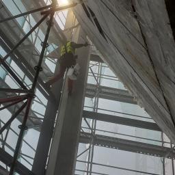 AlpinZerg Sp.z o.o - Pracownik w kamizelce odblaskowej na rusztowaniu, z wiadrem w dłoni, aplikuje pianę montażową na surową, betonową elewację budynku w trakcie ocieplania, widok z dołu.