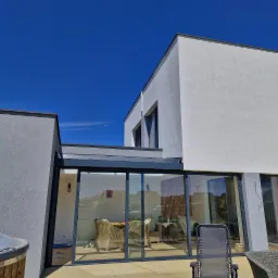 Nowoczesny dom z jasną elewacją i dużymi przesuwnymi oknami tarasowymi, widok z zewnątrz na taras z meblami ogrodowymi, błękitne niebo w tle.