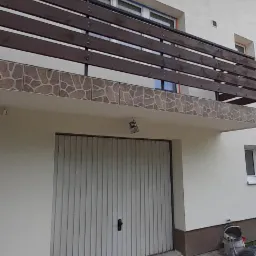 Dwupiętrowy dom z balkonem z brązową drewnianą balustradą i elewacją w kolorze złamanej bieli, widoczne ślady tynkowania na elewacji nad garażem.