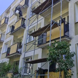 AlpinZerg Sp.z o.o - Pracownik maluje żółte balustrady balkonu na wielopiętrowym budynku, stojąc na podwieszanej platformie z wiadrem i wałkiem, obok rusztowania.