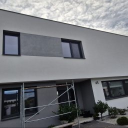 AlpinZerg Sp.z o.o - Nowoczesny dom z dwukolorową elewacją w odcieniach jasnej i ciemnej szarości, z minimalistycznymi czarnymi oknami i numerem 45. Widoczne rusztowanie przy wejściu i osoba z rowerem na podjeździe.