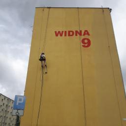 AlpinZerg Sp.z o.o - Pracownik alpinistyczny na linach czyści elewację żółtego bloku z napisem 'WIDNA 9', widoczny znak parkingu dla niepełnosprawnych.