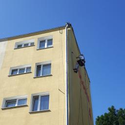 AlpinZerg Sp.z o.o - Pracownik na linie alpinistycznej, w kasku, maluje elewację budynku o żółtym odcieniu, widoczne okna i fragment zieleni w tle, błękitne niebo.