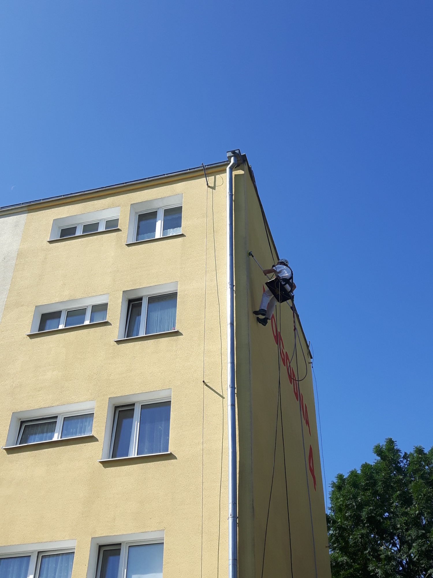 Pracownik na linie alpinistycznej, w kasku, maluje elewację budynku o żółtym odcieniu, widoczne okna i fragment zieleni w tle, błękitne niebo.