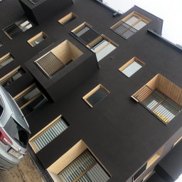 Group - Nowoczesny budynek z ciemną elewacją, licznymi oknami o drewnianych ramach i wystającymi balkonami, widziany z dołu pod kątem, z zaparkowanym samochodem osobowym w lewym dolnym rogu.