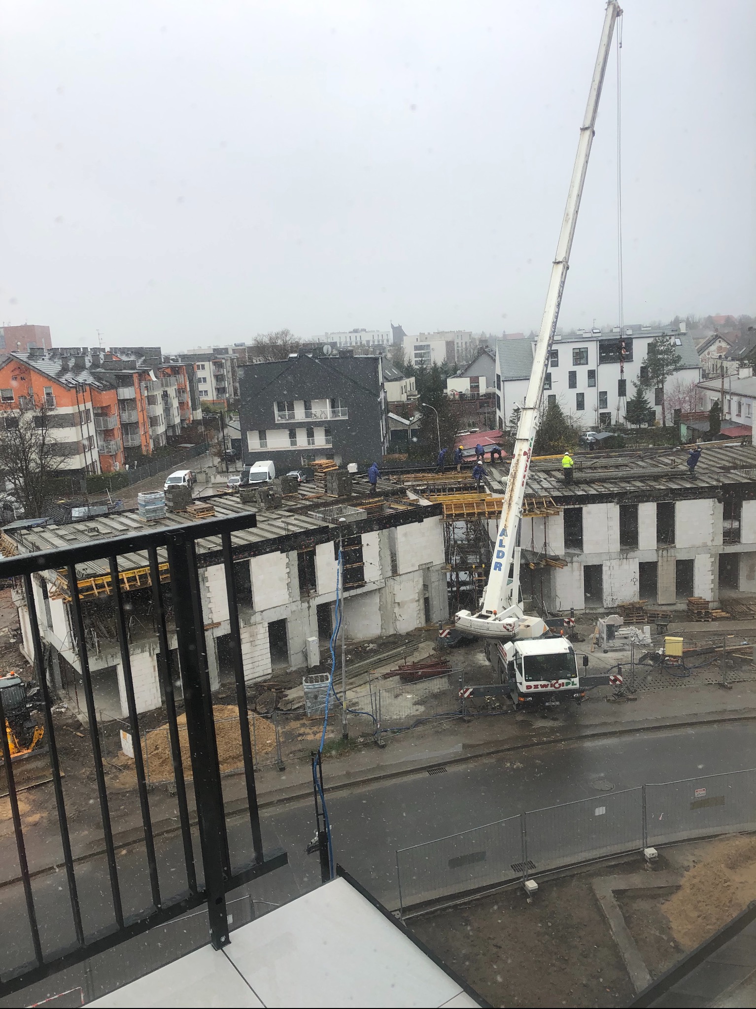 Wysoki dźwig budowlany na placu budowy wielorodzinnego budynku w trakcie wznoszenia, widok z góry w pochmurny dzień, z widocznymi pracownikami na dachu i elementami konstrukcyjnymi.