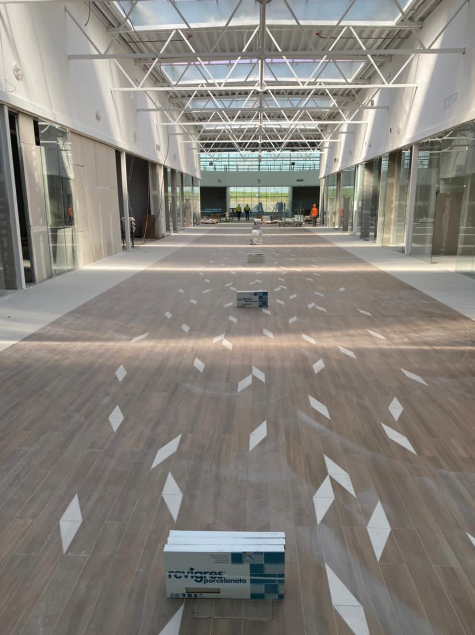 Wnętrze budynku w trakcie wykańczania: ułożona podłoga z paneli z wzorem rombów, widoczne pakiety paneli, szkielet dachu i pracownicy w oddali.