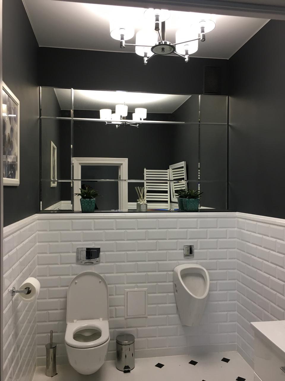 Wnętrze łazienki w stylu nowoczesnym z ciemnymi ścianami i białymi płytkami, duże lustro podzielone na segmenty odbija oświetlenie sufitowe i dekoracje, widoczna toaleta i pisuar.
