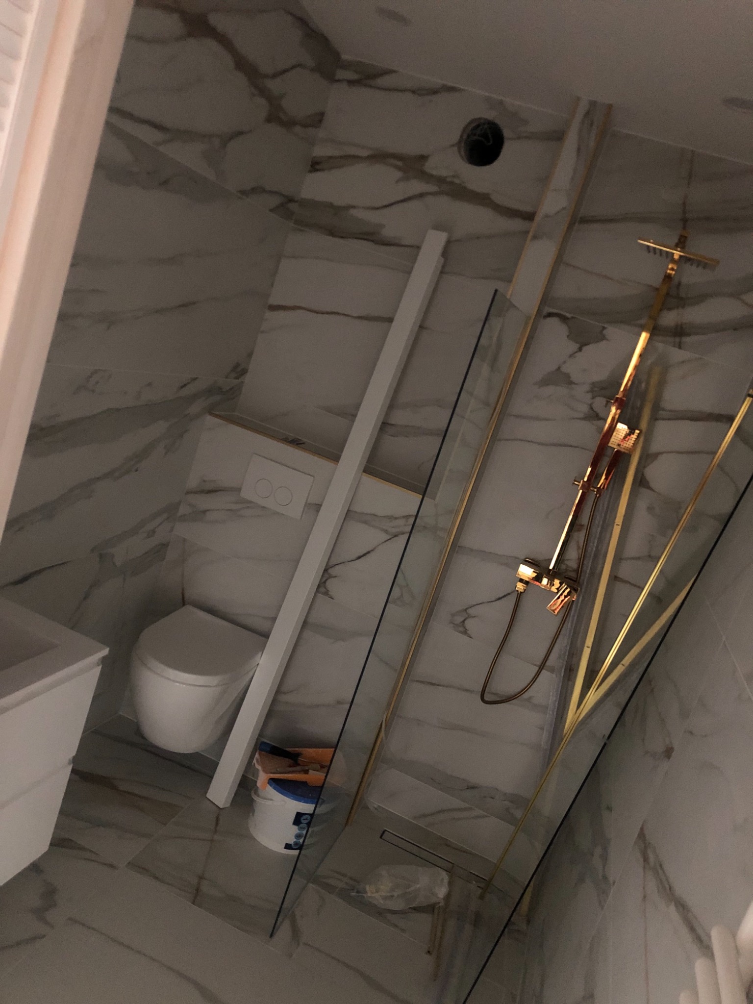 Remont łazienki w trakcie: białe marmurowe płytki, toaleta, umywalka, złota armatura prysznicowa za szklaną kabiną, wiadro z narzędziami malarskimi przy ścianie.