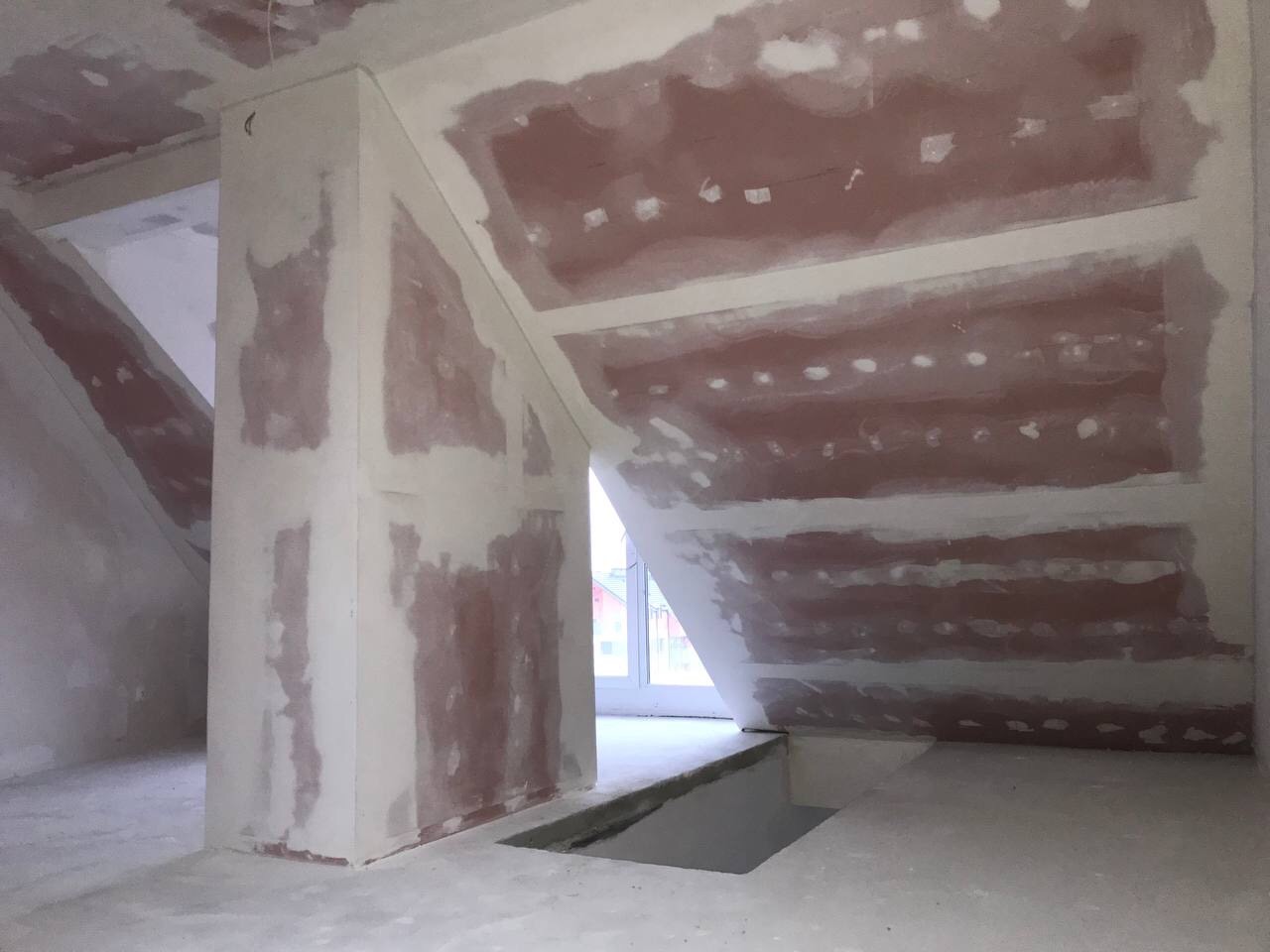 Poddasze w trakcie prac wykończeniowych: ściany i sufit pokryte częściowo nałożoną masą szpachlową, widoczne łączenia płyt kartonowo-gipsowych, otwór w podłodze w pobliżu okna.