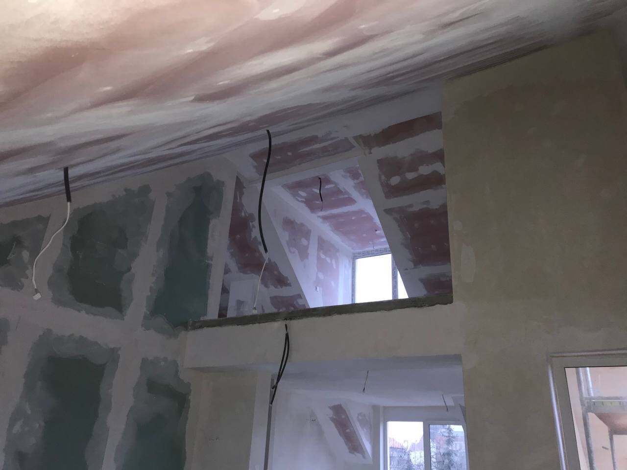 Wnętrze w trakcie prac wykończeniowych: ściany z widocznymi łączeniami płyt gipsowo-kartonowych przed szpachlowaniem, wystające przewody elektryczne, sufit wstępnie wykończony, widok na kolejne...