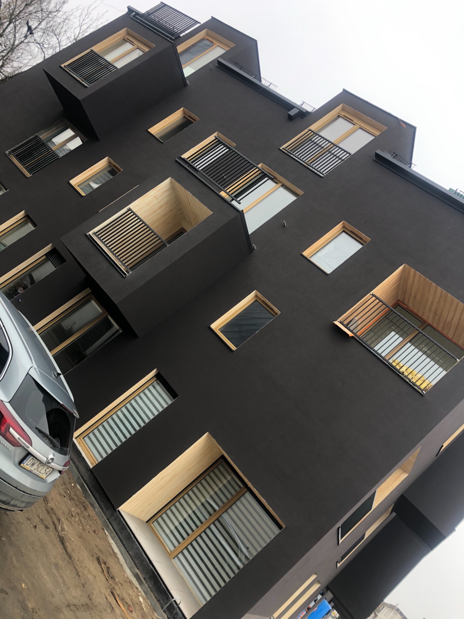 Nowoczesny budynek z ciemną elewacją, licznymi oknami o drewnianych ramach i wystającymi balkonami, widziany z dołu pod kątem, z zaparkowanym samochodem osobowym w lewym dolnym rogu.
