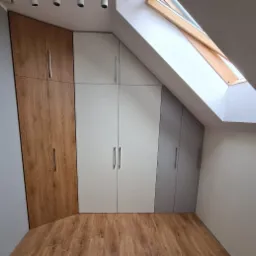 Zabudowa szaf wnękowych z frontami w odcieniach drewna, bieli i szarości, dopasowana do skosu poddasza z oknem dachowym.