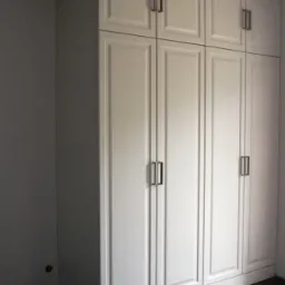 Biała, wysoka szafa z sześcioma drzwiami i ciemnymi, prostokątnymi uchwytami, dopasowana do wnętrza z ciemną podłogą.