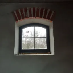 Surowa ściana z szarym tynkiem w trakcie prac wykończeniowych, okno z ceglanym łukiem nadproża, widok na drzewa za oknem.