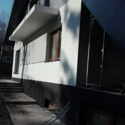 Dwukolorowa elewacja domu jednorodzinnego z widocznym fragmentem dachu i schodami wejściowymi, szare tynki i czarny cokół, widoczne rusztowanie.