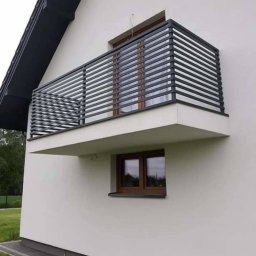 METALSYSTEM Paweł Brzeziński - Nowoczesna balustrada balkonowa z poziomych, grafitowych profili na tle białej elewacji domu z brązową stolarką okienną, z widocznymi workami nawozu u podstawy ściany.