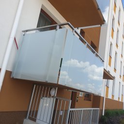 METALSYSTEM Paweł Brzeziński - Balkon z szklaną balustradą i metalową ramą, odbijający błękitne niebo z chmurami, widoczny z dołu, z fragmentem metalowego ogrodzenia przed wejściem do budynku.