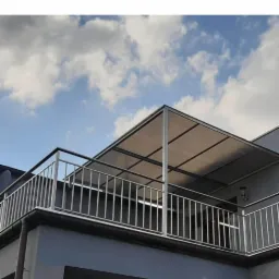 Balkon z metalową balustradą i zadaszeniem z poliwęglanu na tle błękitnego nieba z chmurami.