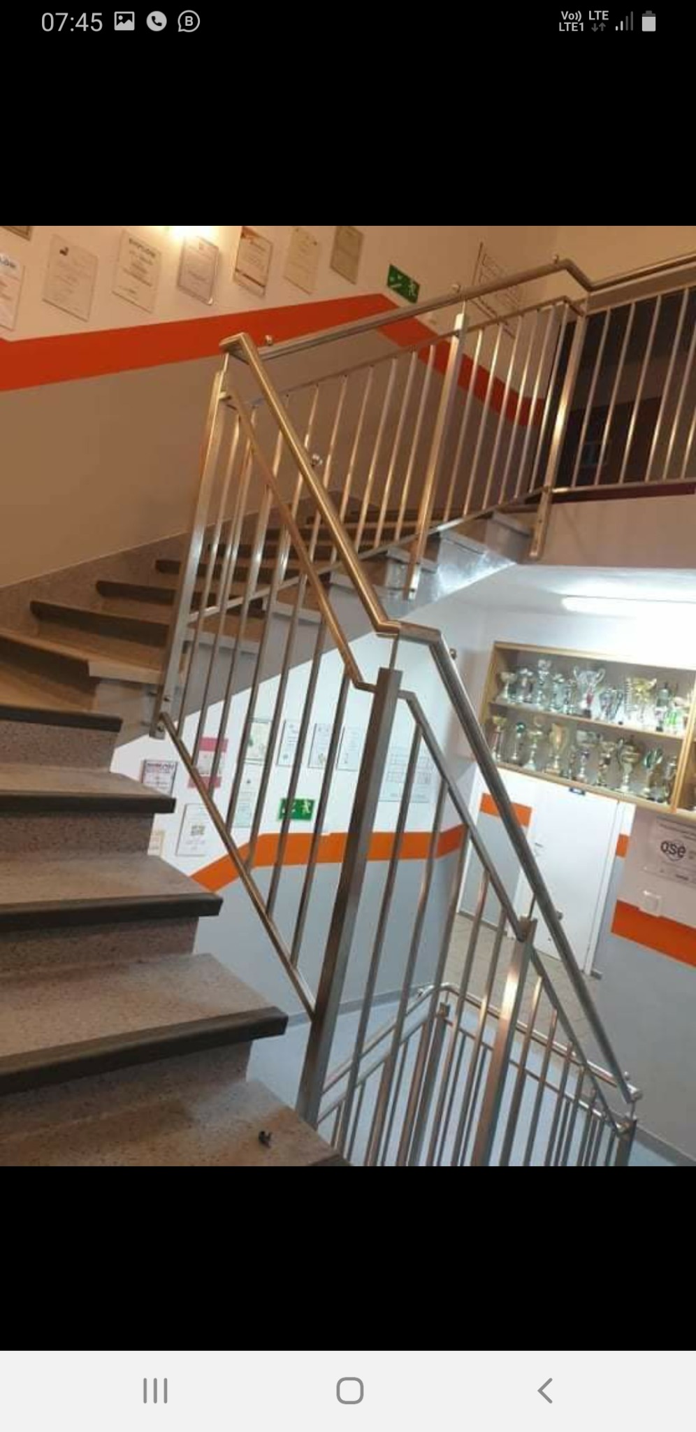 Schody ze stalową balustradą w budynku w Dąbrównie, widoczne certyfikaty na ścianie i półka z pucharami.