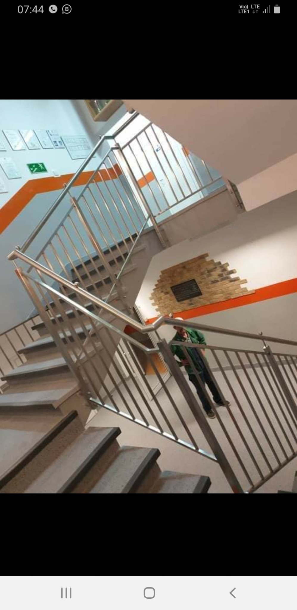 Klatka schodowa z metalową balustradą, pomalowana na biało z pomarańczowymi pasami, widoczna osoba w zielonej kurtce przy poręczy.