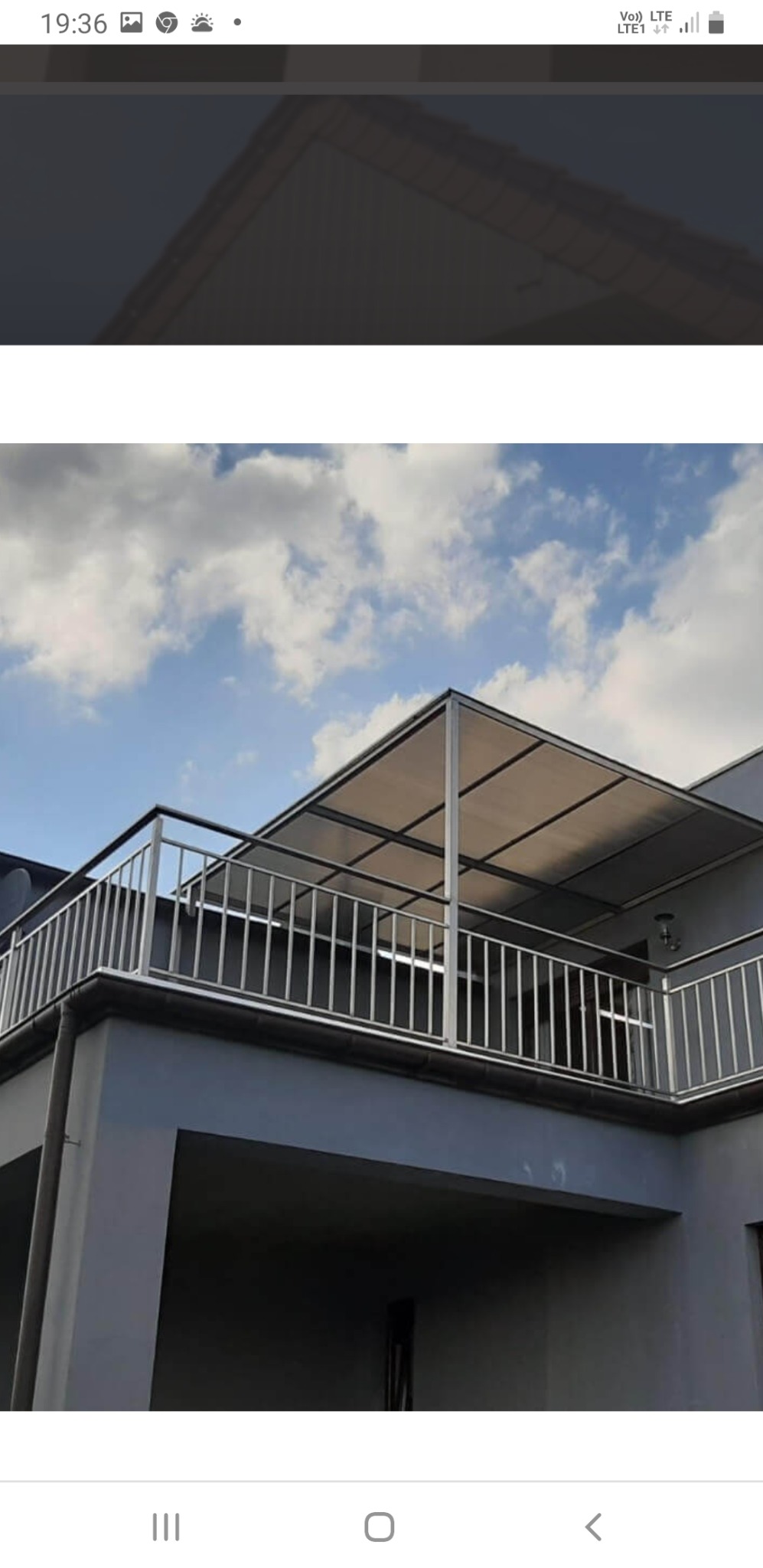 Balkon z metalową balustradą i zadaszeniem z poliwęglanu na tle błękitnego nieba z chmurami.