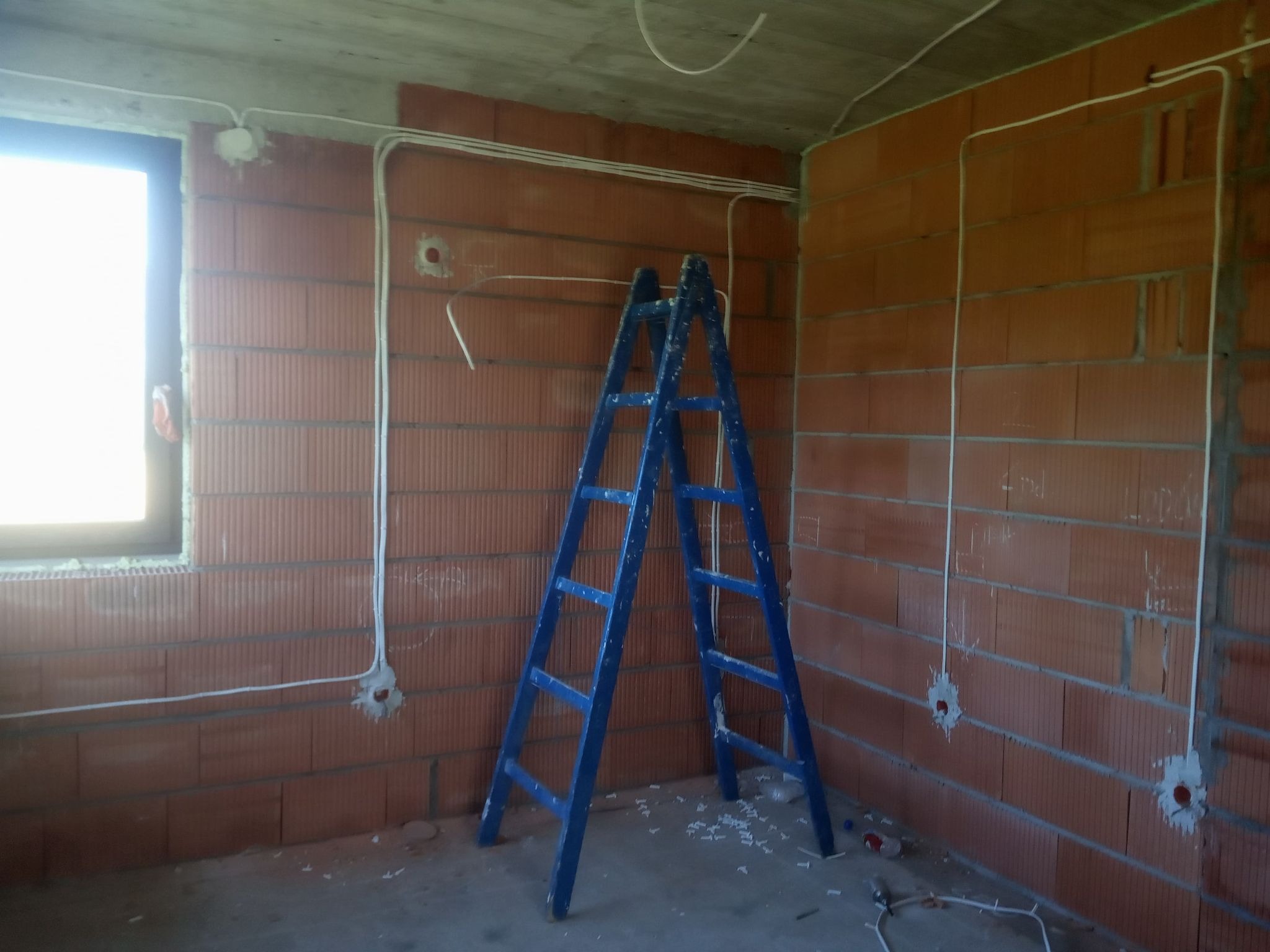 Wnętrze w stanie surowym z widocznymi przewodami elektrycznymi prowadzonymi po ścianach z cegły, niebieska drabina oparta o ścianę, okno w lewym górnym rogu.