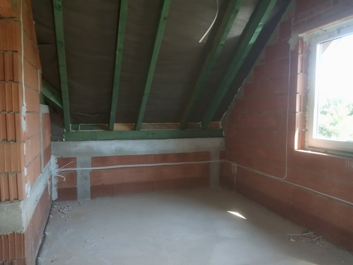 Surowy stan poddasza z widocznymi przewodami elektrycznymi poprowadzonymi wzdłuż ceglanej ściany przed tynkowaniem, drewniana konstrukcja dachu i okno w tle.