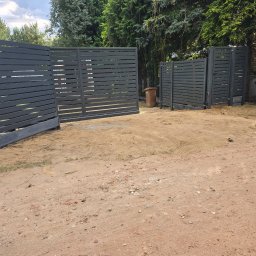 P. K. J ZAUNE Piotr Prokurat - Nowoczesna, pozioma brama wjazdowa z szarego metalu, z widocznymi szczelinami, na tle zieleni drzew. Brama otwarta, wjazd na żwirową posesję.