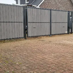 Nowoczesna brama wjazdowa z szarego kompozytu imitującego drewno, osadzona w czarnej metalowej ramie z numerem posesji '5', na tle brukowanej kostki i domu z czerwonej cegły.