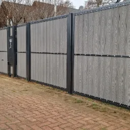 Nowoczesne ogrodzenie posesji z szarych paneli imitujących drewno, z czarną metalową ramą i widoczną furtką z numerem domu '5'.