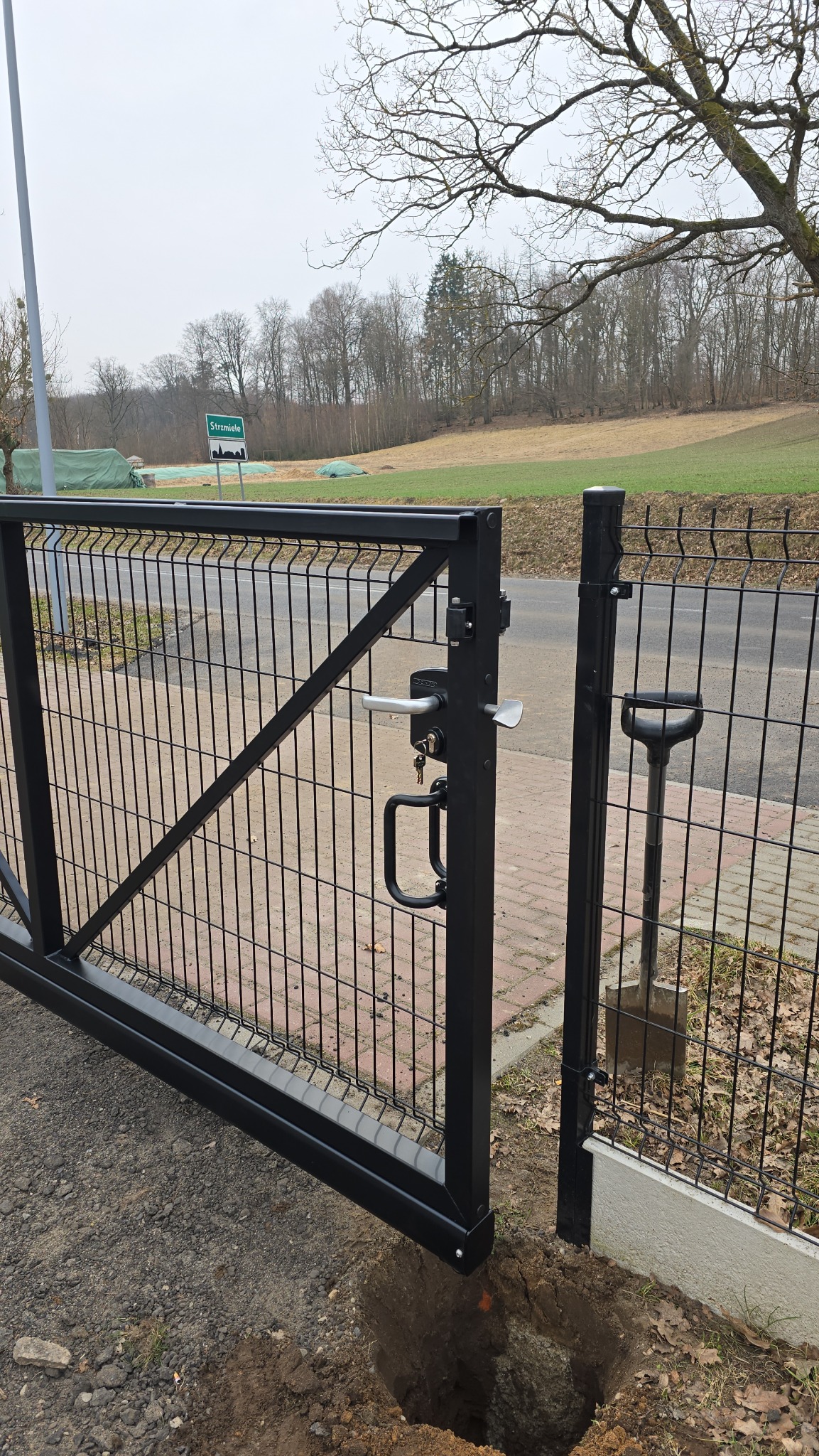 Otwarta, czarna furtka z siatki panelowej, z widoczną klamką i zamkiem. W tle zielony krajobraz i znak drogowy. Obok furtki wbita łopata.