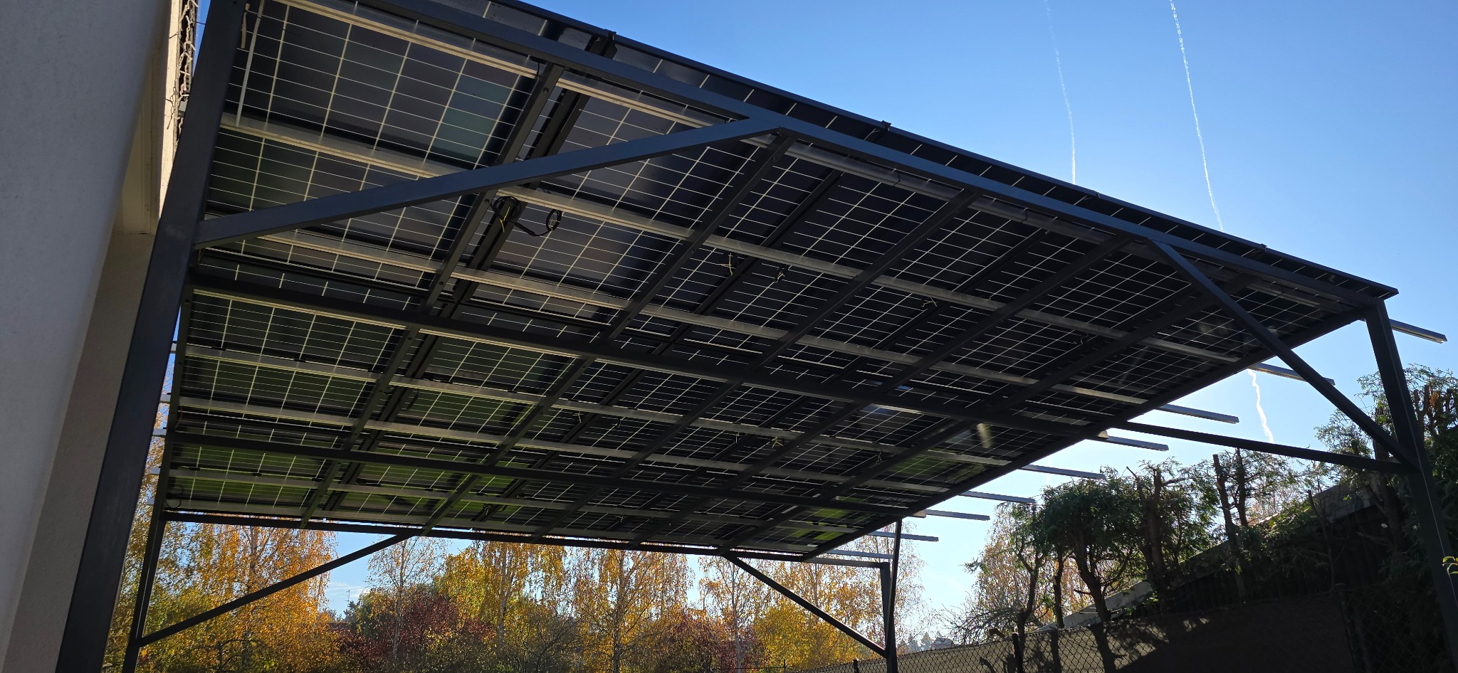 Metalowa konstrukcja z panelami fotowoltaicznymi, widok od spodu, z widocznym niebem i drzewami w tle. Konstrukcja pomalowana na czarno, panele w kratkę.
