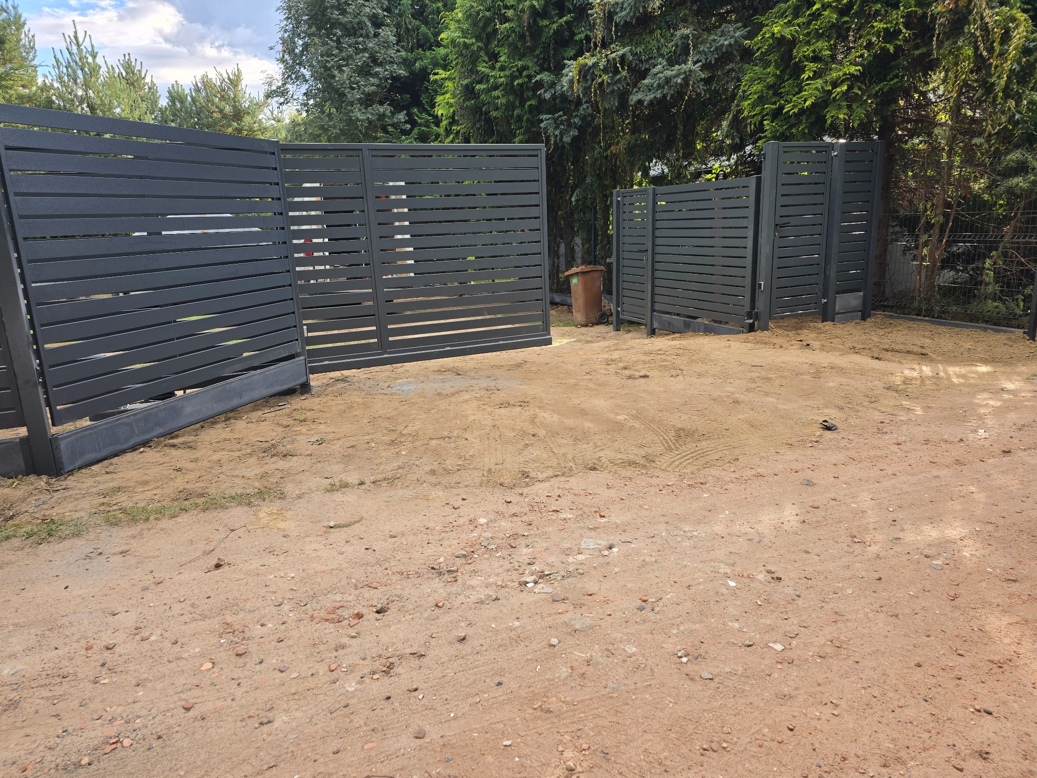 Nowoczesna, pozioma brama wjazdowa z szarego metalu, z widocznymi szczelinami, na tle zieleni drzew. Brama otwarta, wjazd na żwirową posesję.