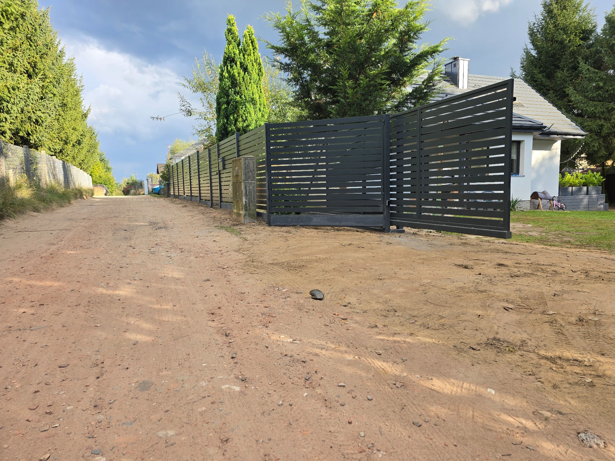 Nowoczesna, pozioma, metalowa brama wjazdowa w kolorze grafitowym, z widocznym fragmentem ogrodzenia. Brama otwarta, na tle domu i zieleni. Ujęcie z poziomu gruntu.